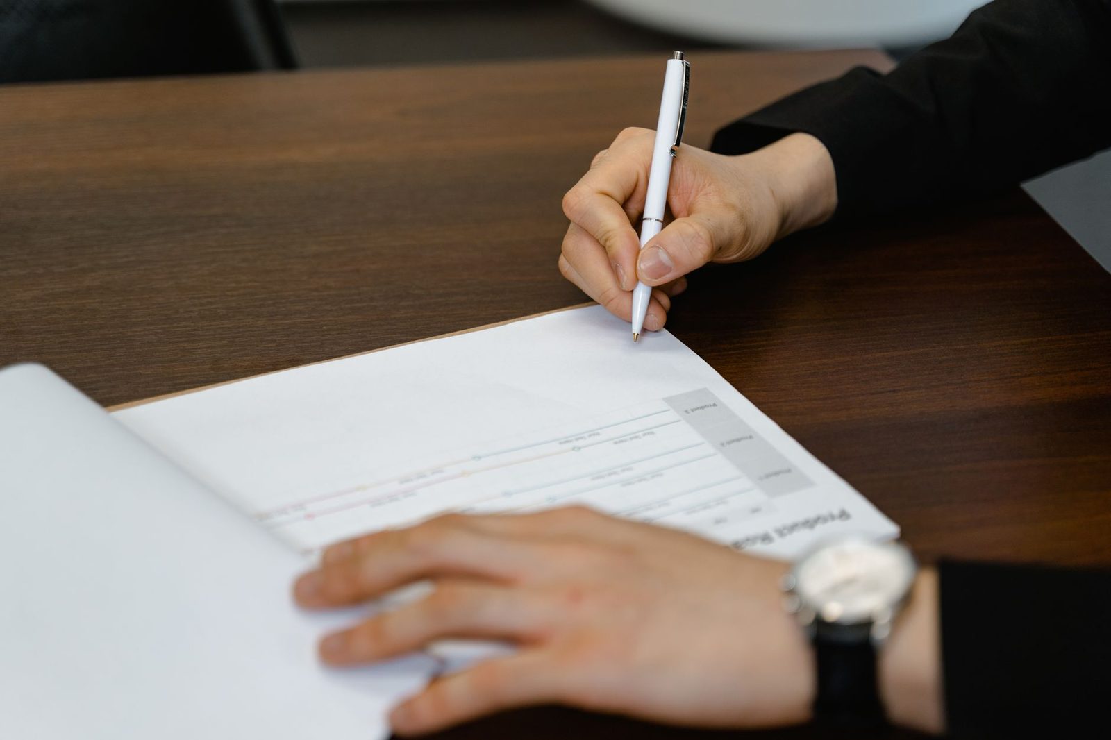 A hand with a white pen over a printed sheet on a dark wooden desk