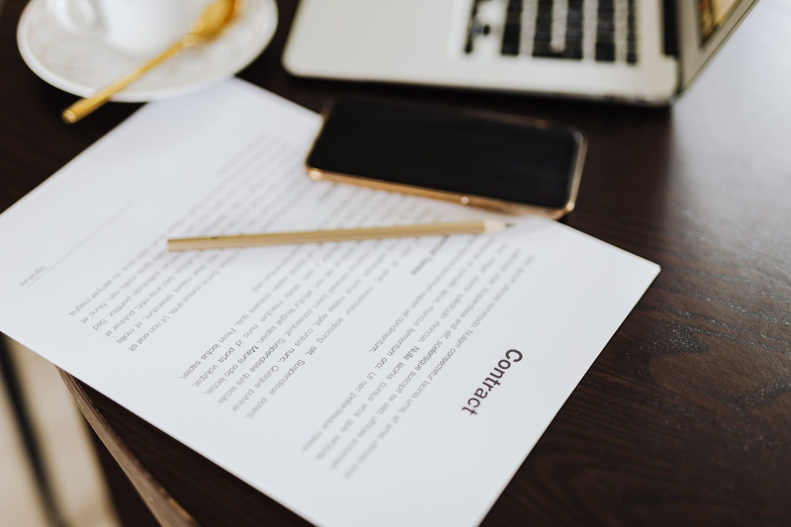 A printed contract with a gold pencil, a phone and a laptop on a dark wooden desk